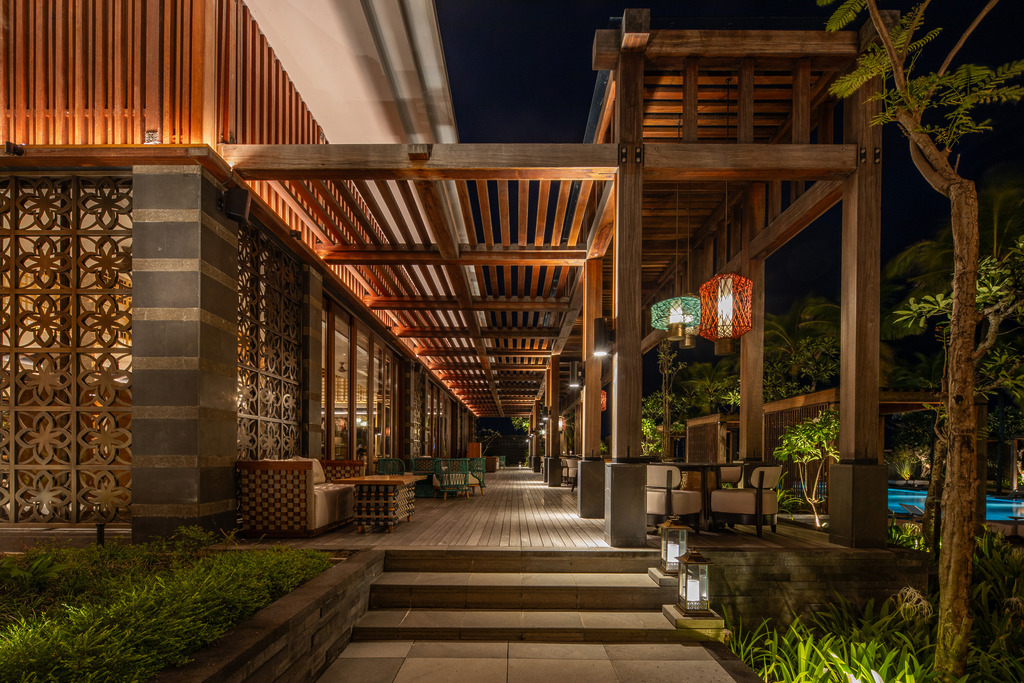Regent Bali Canggu &mdash; a night view along a warmly lit timber pergola walkway with carved stone fretwork walls, coloured pendant lanterns, and a glimpse of the illuminated pool beyond.