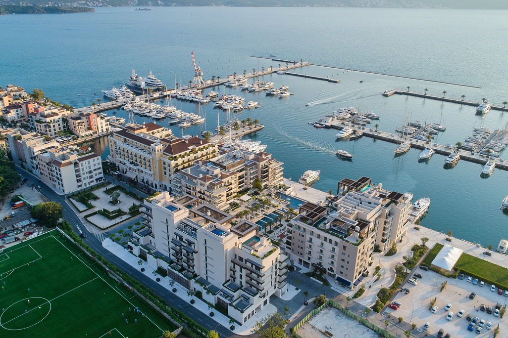 Aerial view of Porto Montenegro resort and marina with superyachts, residential blocks, rooftop pools, and a football pitch in the foreground, Montenegro