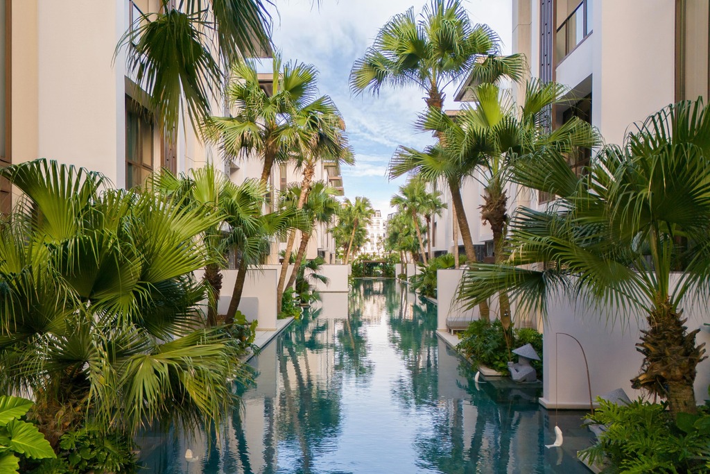 Lush resort poolscape flanked by palm trees and low-rise residence buildings with warm stone facades, InterContinental Halong Bay by WATG
