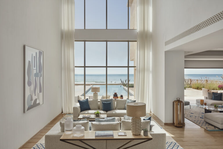 High ceiling white living room with glass panels walls and white sofa with blue cushions overseeing the pool and sea