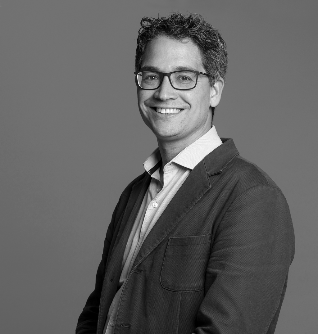 Black and white portrait of Tony Menezes, WATG, smiling against a grey backdrop.