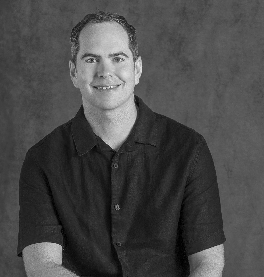 Black and white portrait of Guy Cooke, Associate Principal of Advisory Services, WATG, smiling against a grey backdrop.