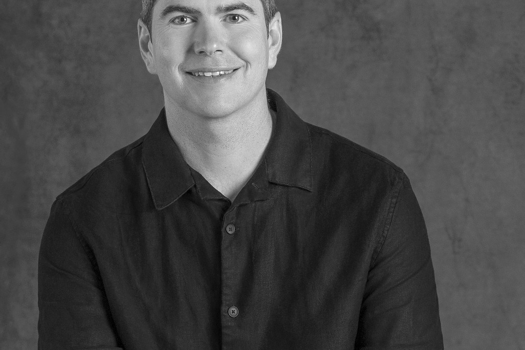 Black and white portrait of Guy Cooke, Associate Principal of Advisory Services, WATG, smiling against a grey backdrop.
