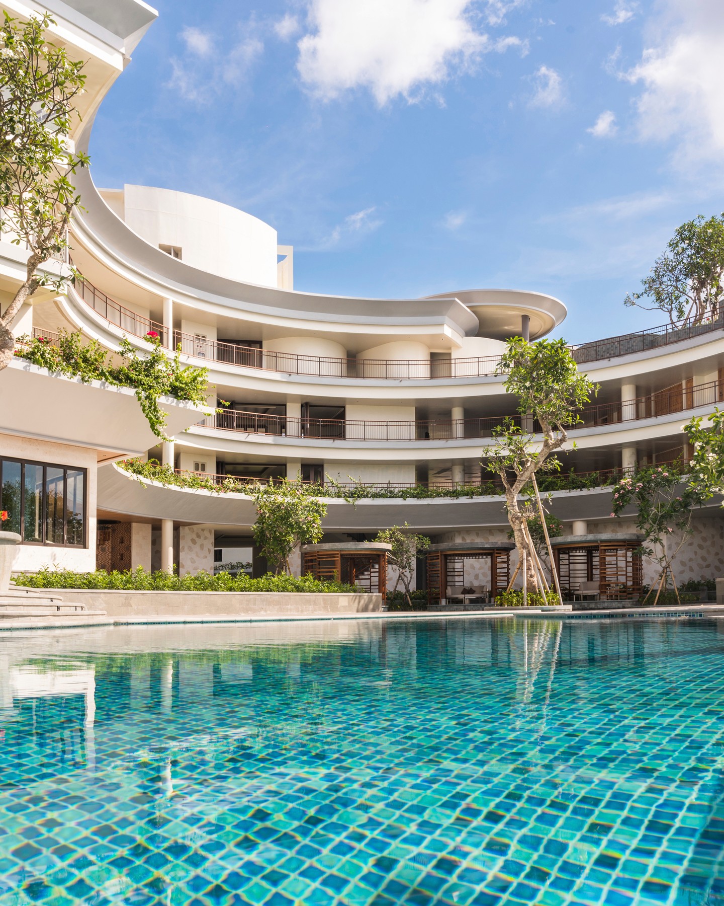 A teal pool with mosaic tile sits in front of a curved hotel balcony with palm trees