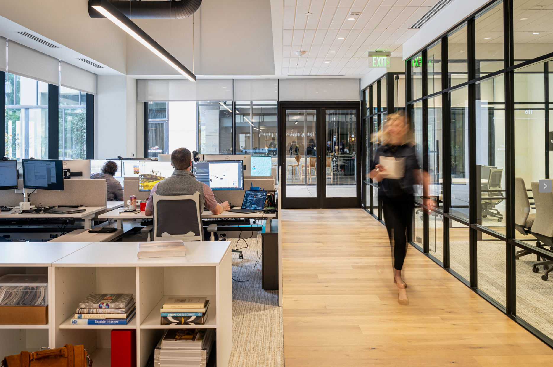 WATG Dallas office open-plan workspace with staff at multi-monitor desks, glass-partitioned meeting rooms, and timber flooring.