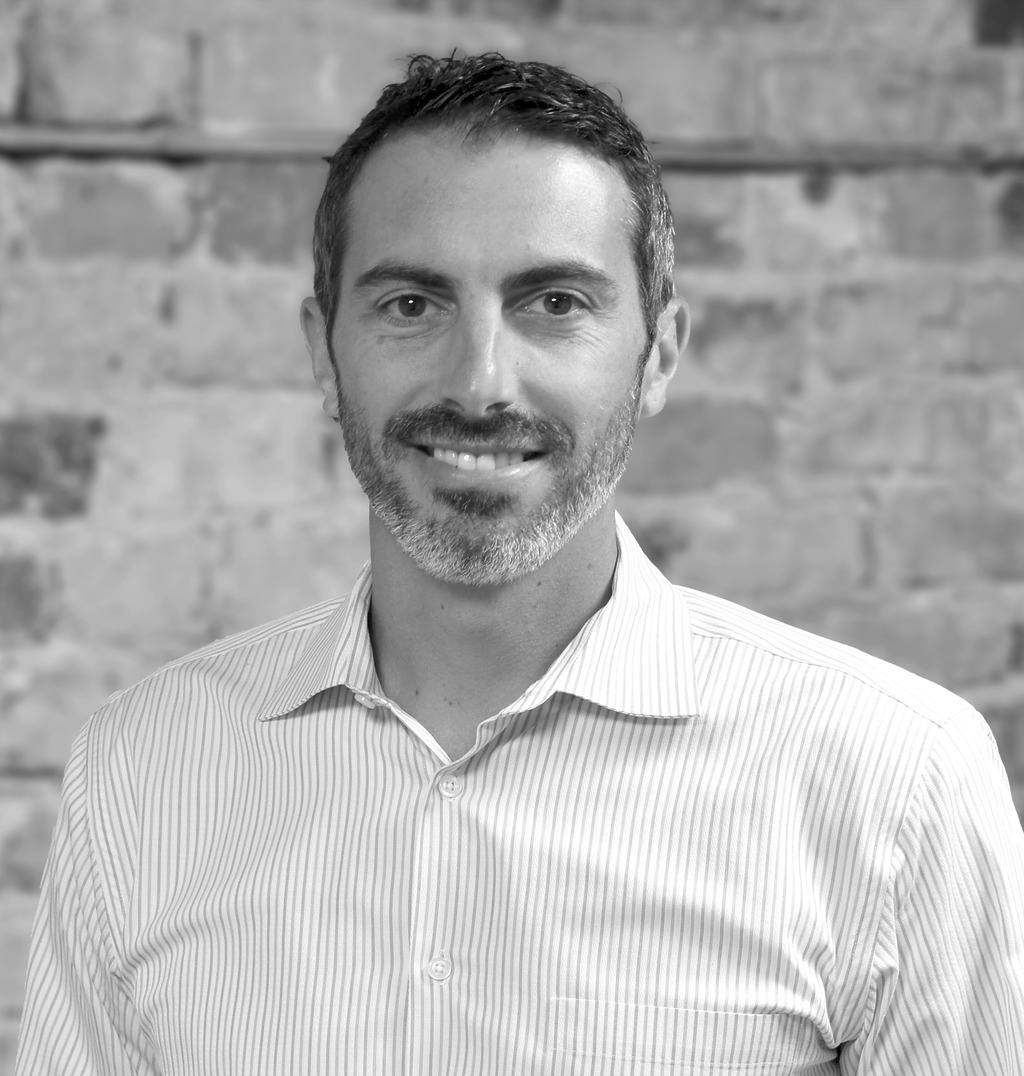 Black and white portrait of John De Richie III , WATG, smiling against a brick wall.