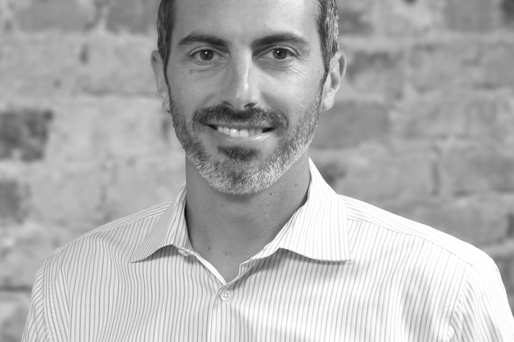 Black and white portrait of John De Richie III , WATG, smiling against a brick wall.