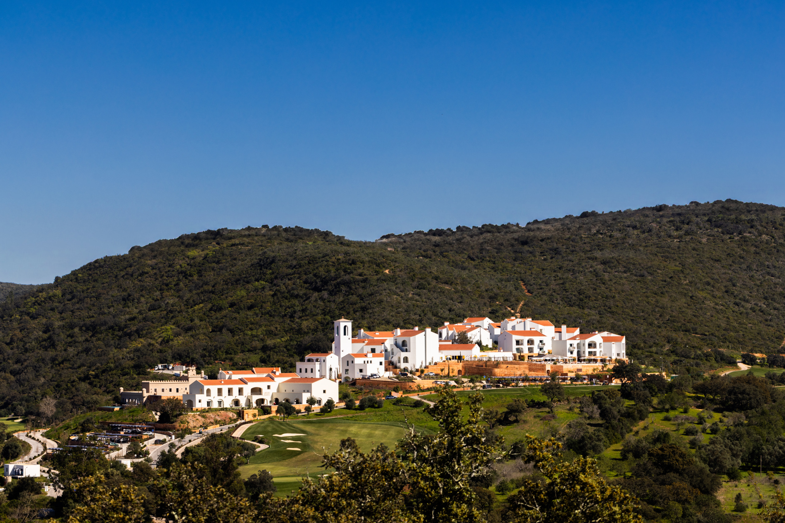 Viceroy at Ombria Algarve, a cluster of whitewashed buildings with terracotta roofs and a bell tower, set on a wooded hillside above a golf course.