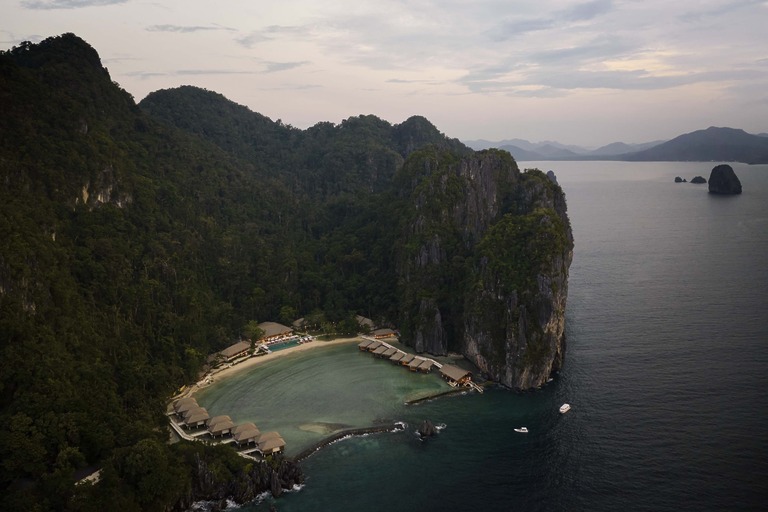 An aerial twilight view of Lagen El Nido showing the resort&rsquo;s integration into a private limestone cove, featuring water cottages and lush tropical forest.