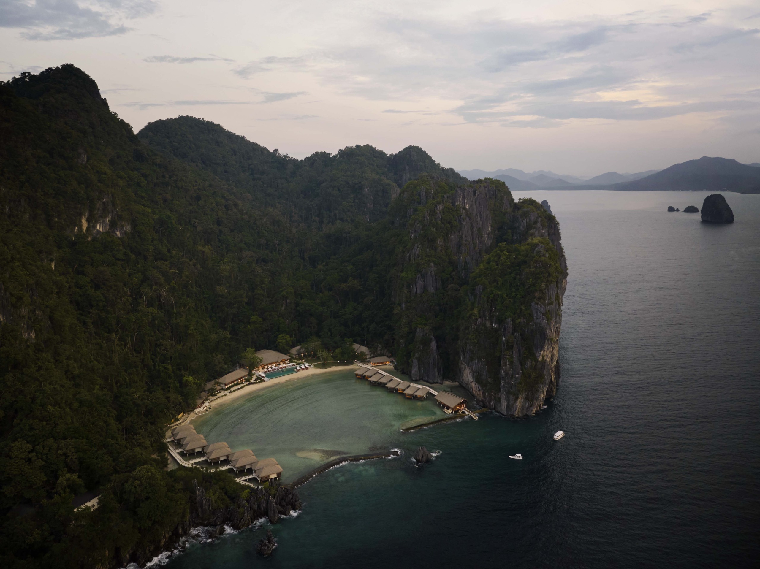 Lagen Island Resort El Nido — aerial dusk view of the entire resort nestled within a sheltered cove, with thatched overwater villas and a white sand beach enclosed by dense jungle-covered limestone cliffs and open sea beyond.