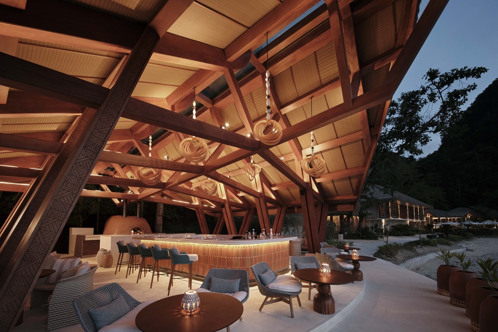 A warm, evening perspective of the beachfront bar highlighting the bespoke lighting design, lounge areas, and a wood-fired oven under a structural canopy.