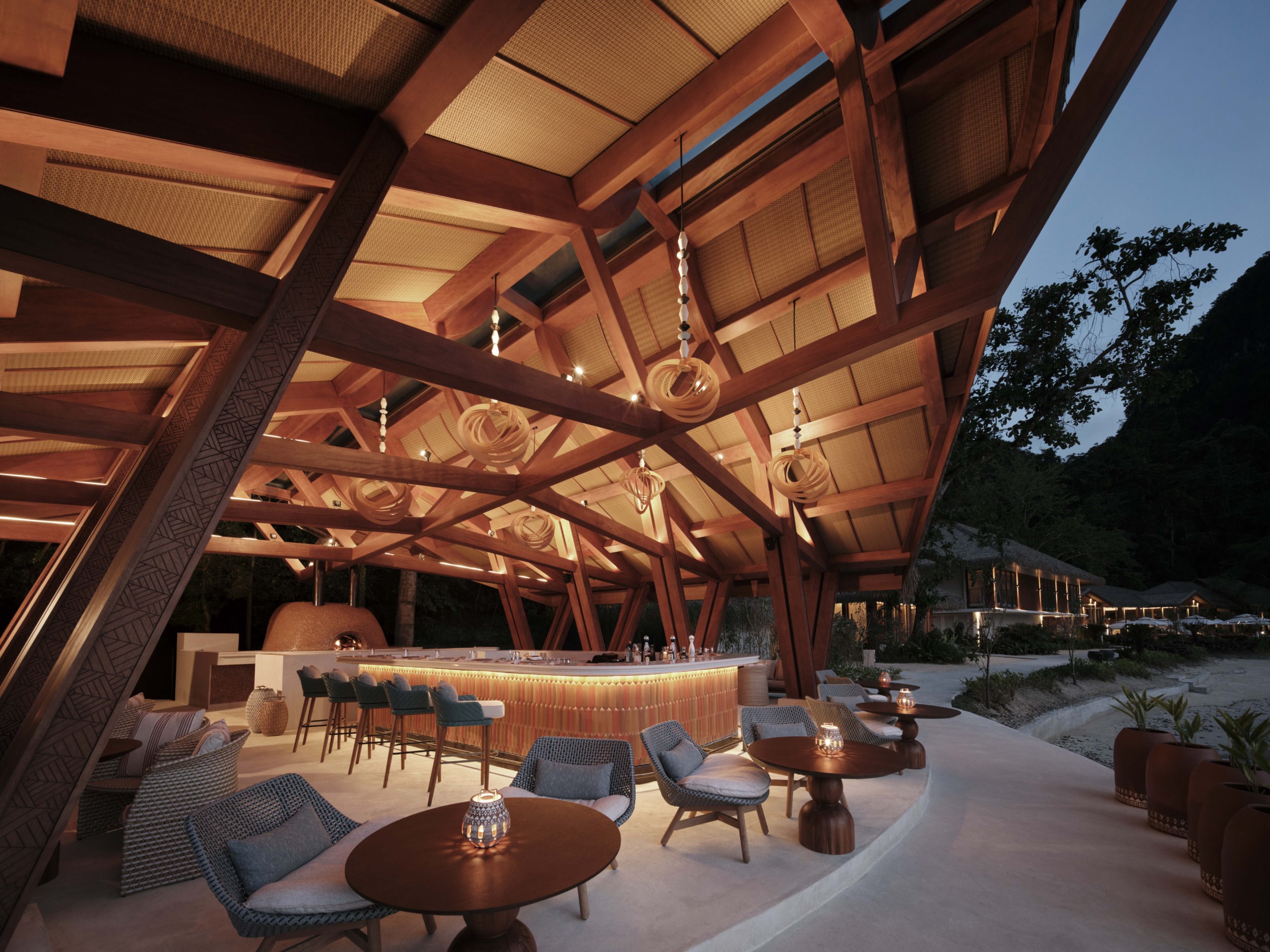 A warm, evening perspective of the beachfront bar highlighting the bespoke lighting design, lounge areas, and a wood-fired oven under a structural canopy.
