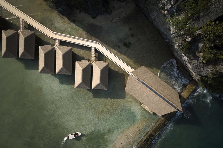 Lagen Island Resort El Nido &mdash; aerial view of a row of thatched overwater villas connected by a timber boardwalk angling from the limestone shoreline into the turquoise shallows, with a small boat passing below.