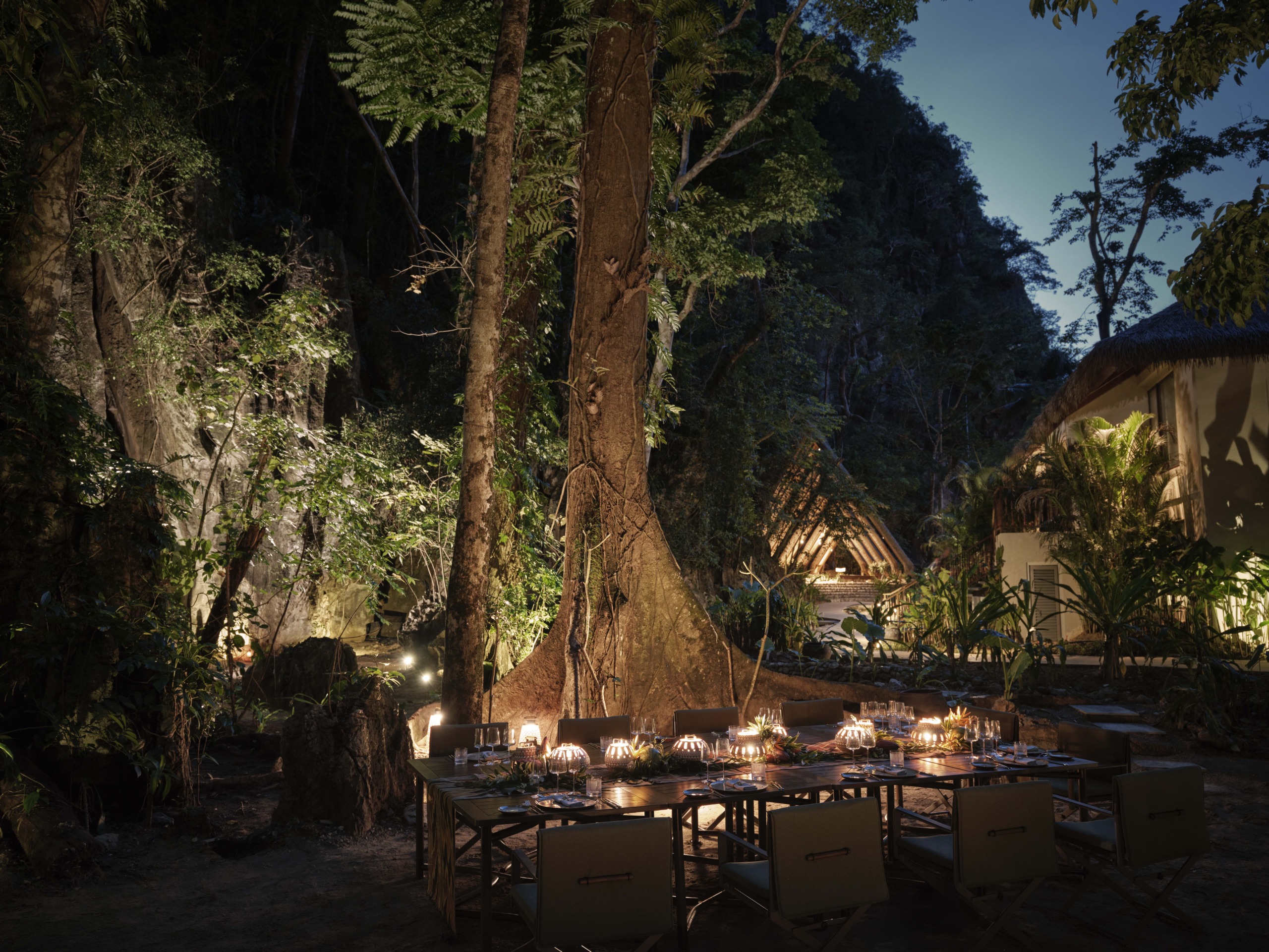 A secluded forest dining setting under the canopy of an ancient tree, illuminated by lanterns to create an intimate atmosphere for events or weddings.