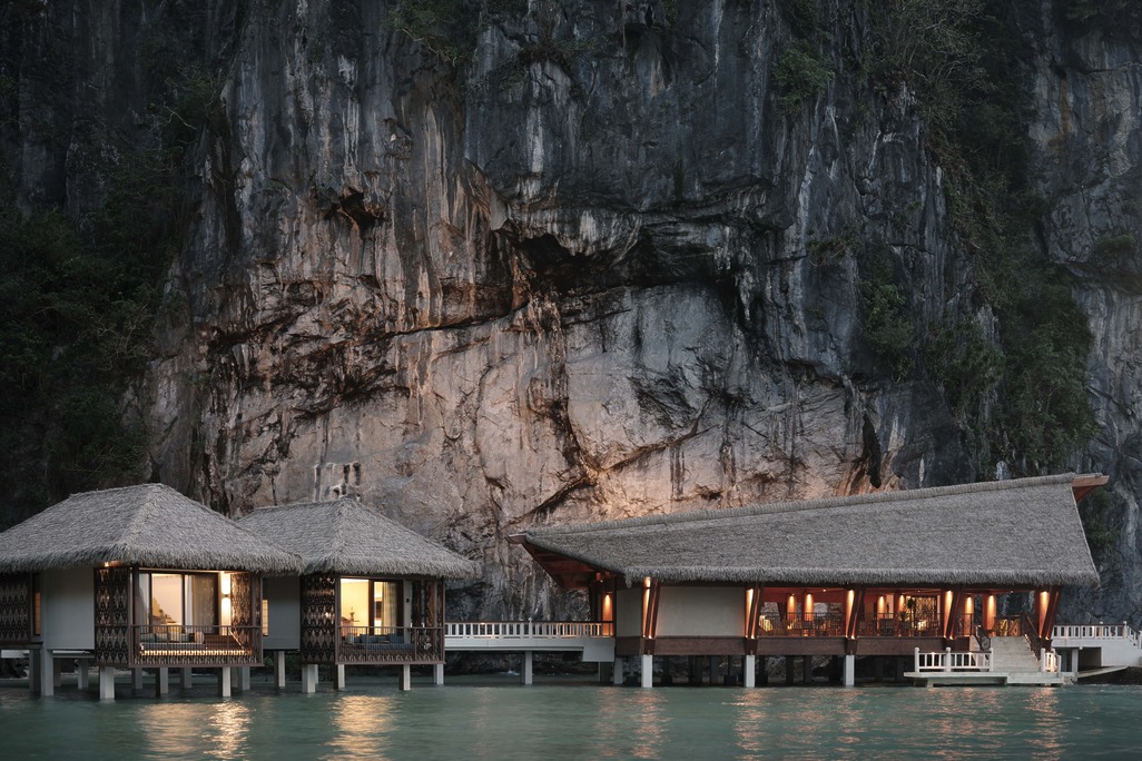 An evening view of the overwater villas and lounge at Lagen El Nido, nestled at the base of a towering, illuminated limestone cliff.