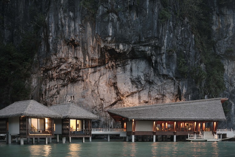 An evening view of the overwater villas and lounge at Lagen El Nido, nestled at the base of a towering, illuminated limestone cliff.