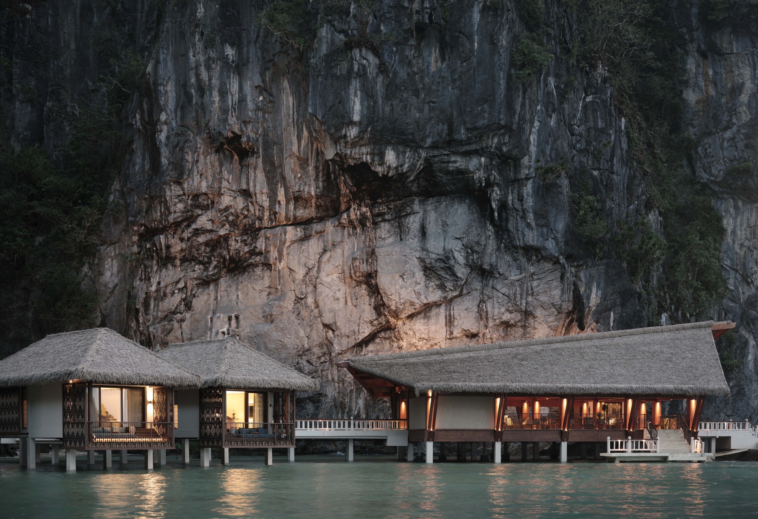 An evening view of the overwater villas and lounge at Lagen El Nido, nestled at the base of a towering, illuminated limestone cliff.