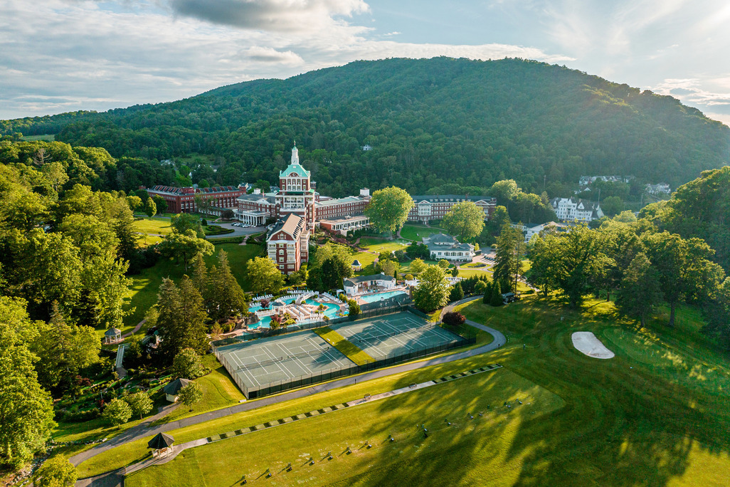 An aerial wide-angle shot of the historic Omni Homestead Resort nestled in the lush, green Allegheny Mountains. The image shows the iconic red-brick tower and wings, manicured tennis courts in the foreground, a swimming pool complex, and the vast rolling greens of the golf course under a bright afternoon sun.