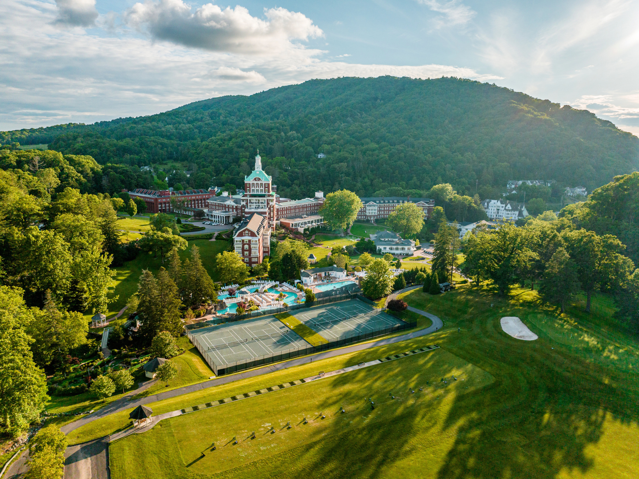 An aerial wide-angle shot of the historic Omni Homestead Resort nestled in the lush, green Allegheny Mountains. The image shows the iconic red-brick tower and wings, manicured tennis courts in the foreground, a swimming pool complex, and the vast rolling greens of the golf course under a bright afternoon sun.