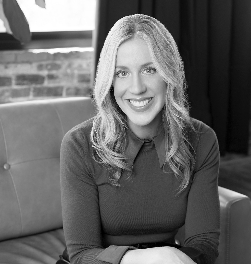 Black and white professional headshot of Molly O'Keefe seated on a leather sofa against an exposed brick wall.