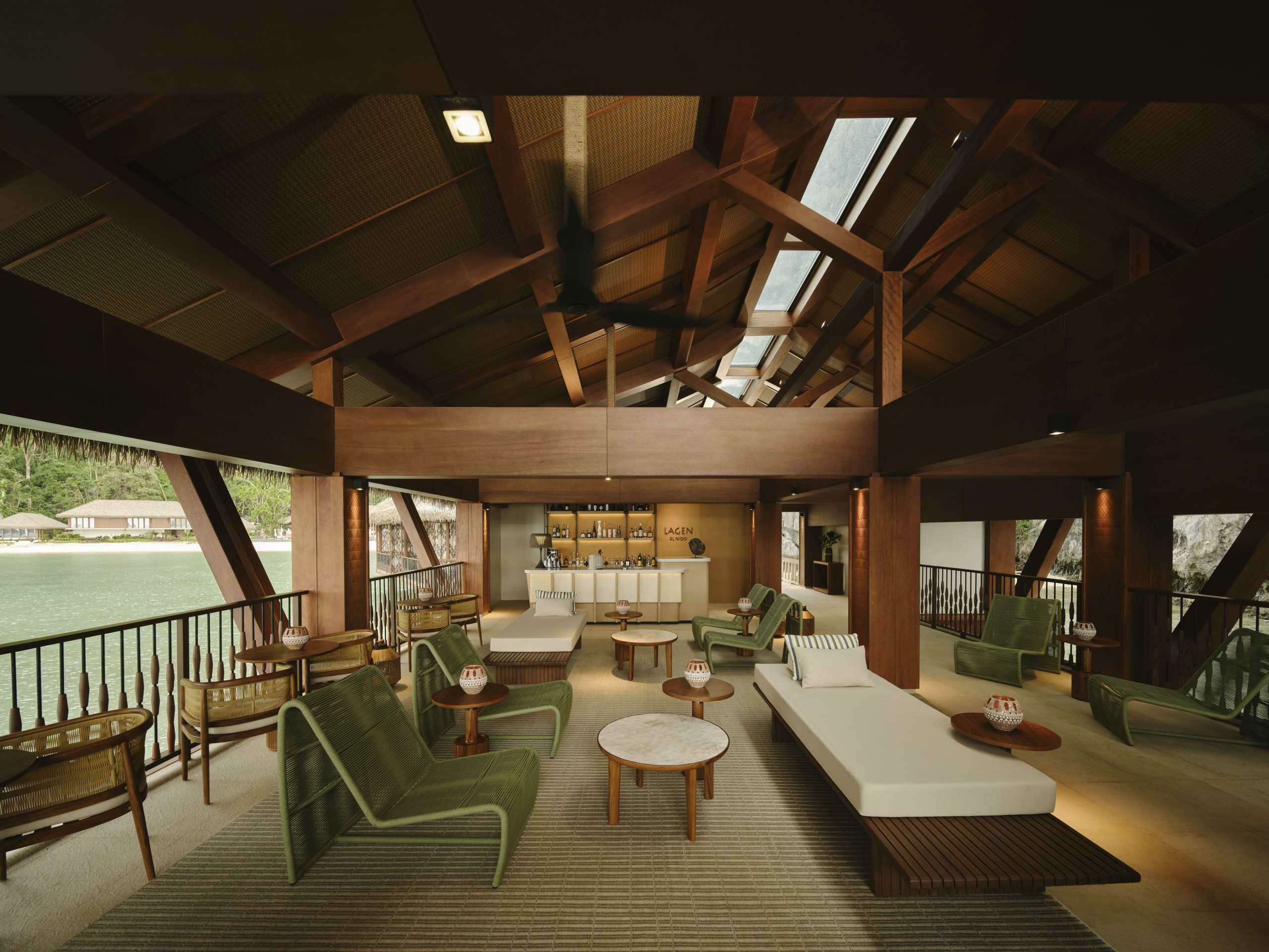 Lagen Island Resort El Nido overwater bar and lounge with exposed timber trusses, green lounge chairs, a daybed, and open views across the bay to the beach.