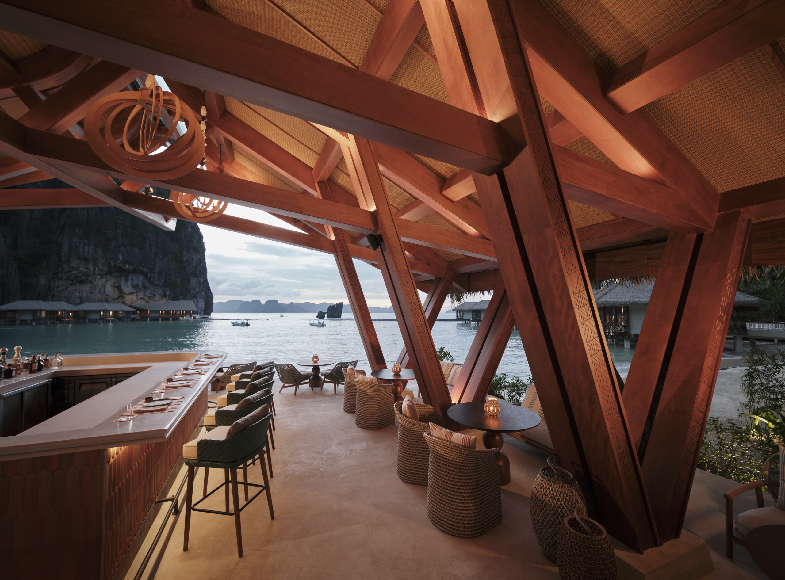 Lagen Island Resort El Nido beachside bar framed by angled carved timber trusses, with bar stools, woven lounge chairs, and panoramic bay and limestone cliff views beyond.