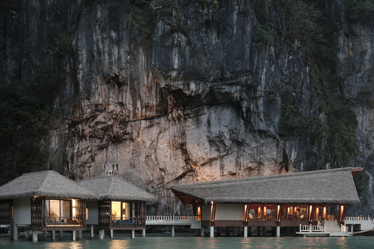 Lagen Island Resort El Nido illuminated overwater bungalows with nipa thatched roofs nestled directly beneath a towering limestone cliff face at dusk.