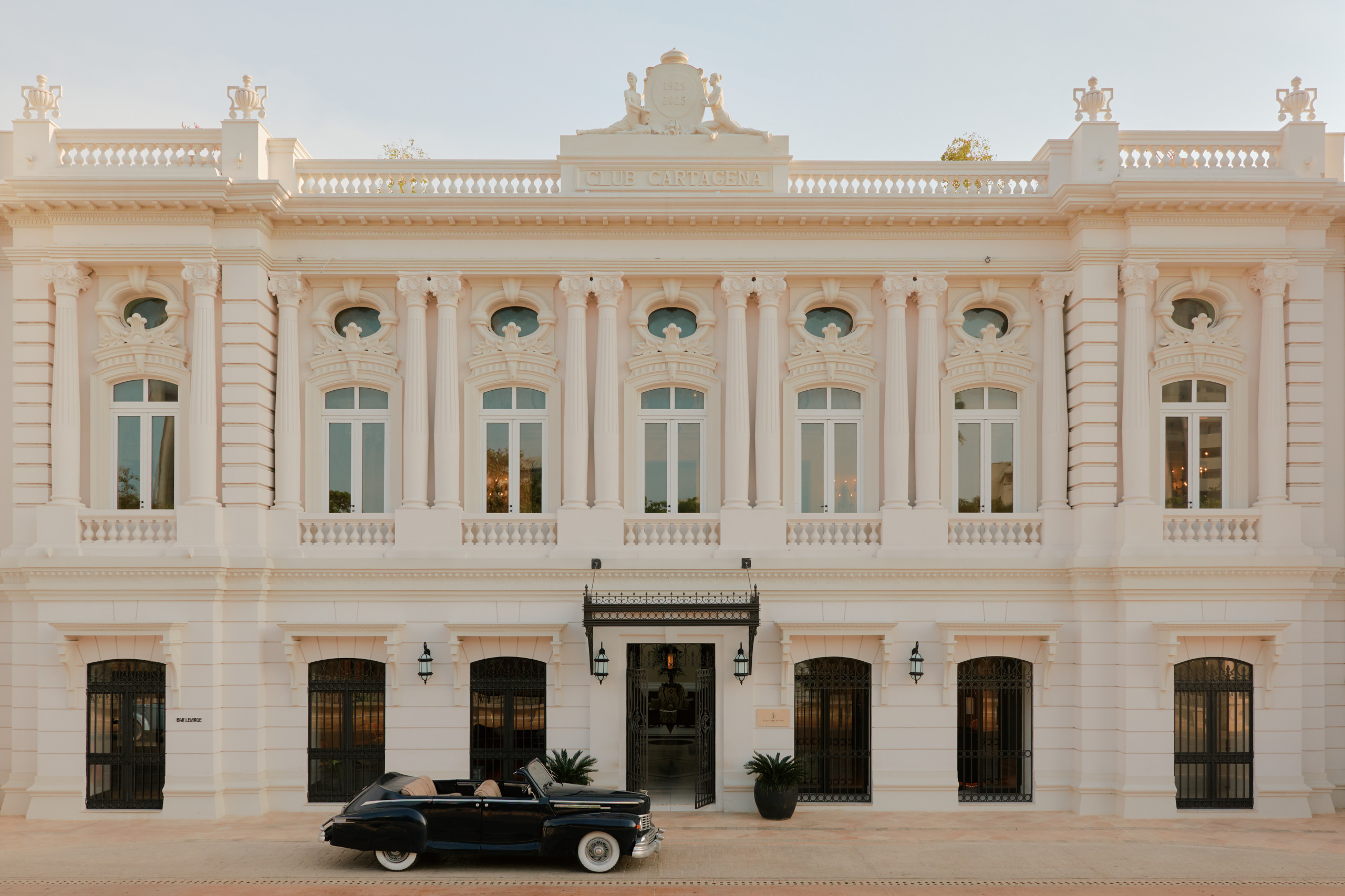 The ornate cream-coloured neoclassical facade of the Four Seasons Hotel Cartagena, Colombia, featuring symmetrical pilasters, arched ground-floor doors with wrought iron grilles, tall first-floor windows with balustrades, oval oculus windows on the upper storey, and a decorative roofline with urns and a central coat-of-arms pediment. A dark navy vintage convertible automobile with white-wall tyres is parked in front of the canopied main entrance, flanked by lantern wall lights and potted palms.