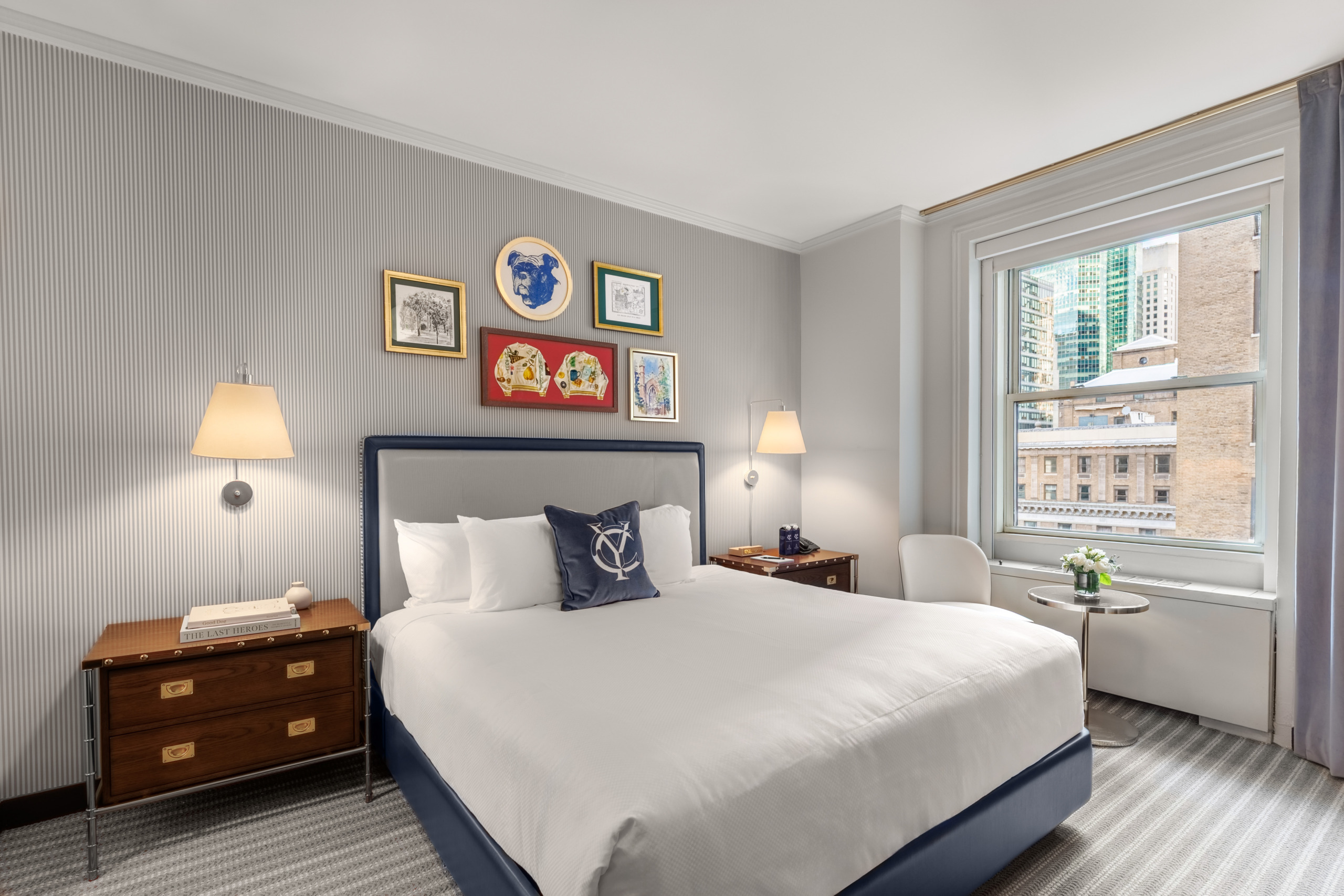 Wide-angle view of a renovated Yale Club of New York City guest room by Wimberly Interiors, showing a navy-framed king bed, campaign nightstands, striped wallpaper, wall-mounted sconces, and a large window overlooking the Midtown Manhattan skyline.