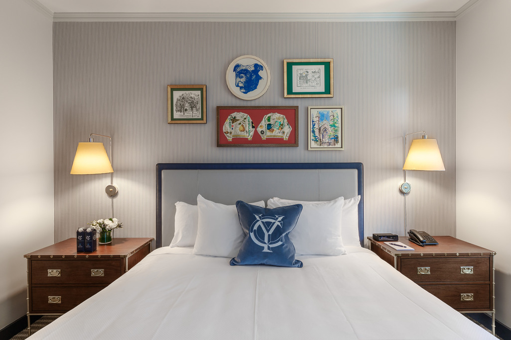 Wimberly Interiors-designed guest room at The Yale Club of New York City featuring a king bed with navy-trimmed upholstered headboard, a blue velvet throw pillow embroidered with the Yale 'YC' monogram, campaign-style walnut nightstands, and a gallery wall of Yale-themed artwork including a bulldog portrait and framed varsity sweaters, set against blue-grey ticking-stripe wallpaper.