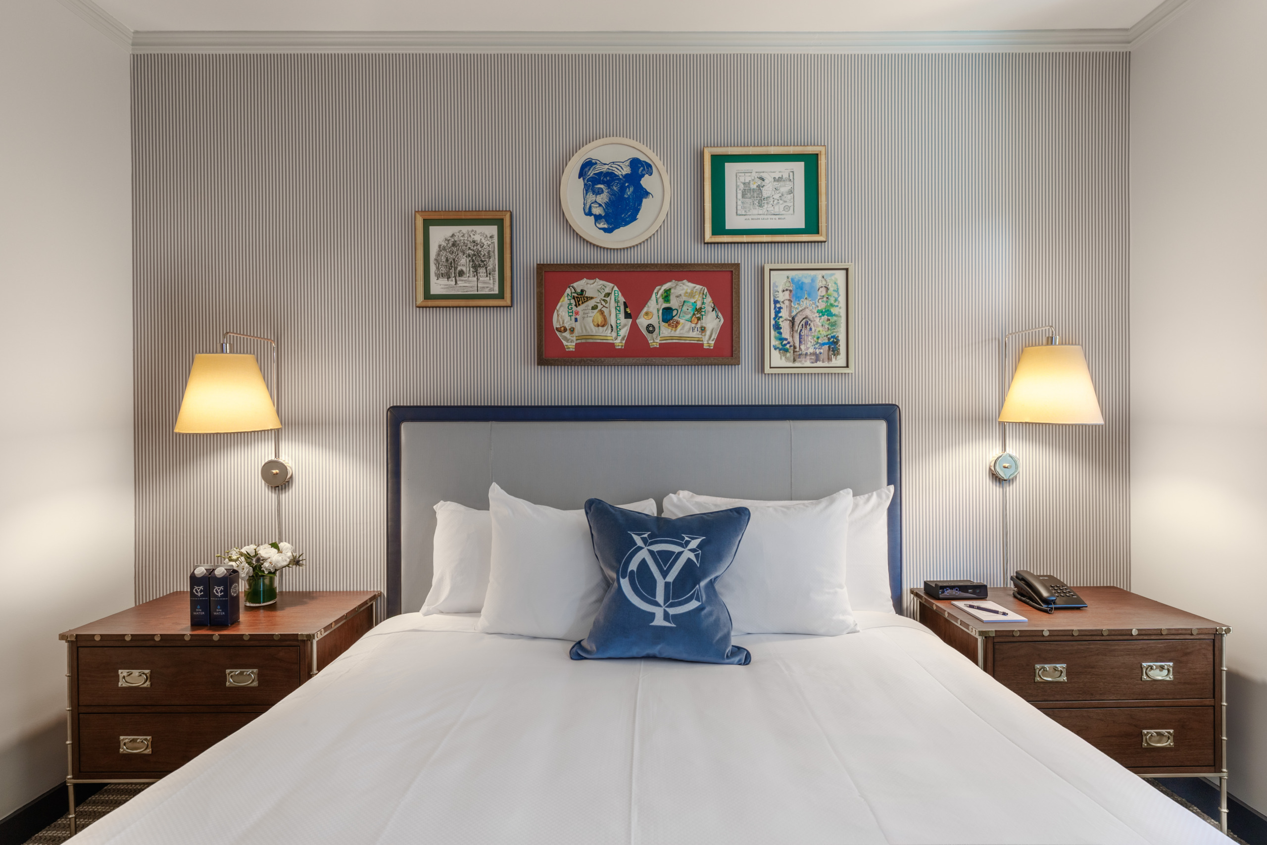 Wimberly Interiors-designed guest room at The Yale Club of New York City featuring a king bed with navy-trimmed upholstered headboard, a blue velvet throw pillow embroidered with the Yale 'YC' monogram, campaign-style walnut nightstands, and a gallery wall of Yale-themed artwork including a bulldog portrait and framed varsity sweaters, set against blue-grey ticking-stripe wallpaper.