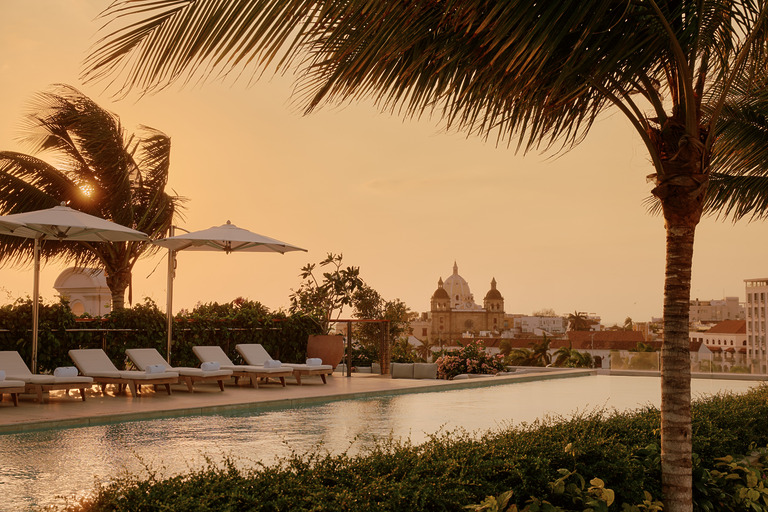 Four Seasons Hotel Cartagena rooftop pool at sunset, with sun loungers and palm trees framing a view over the historic domes and terracotta rooftops of Cartagena's walled city, Colombia.