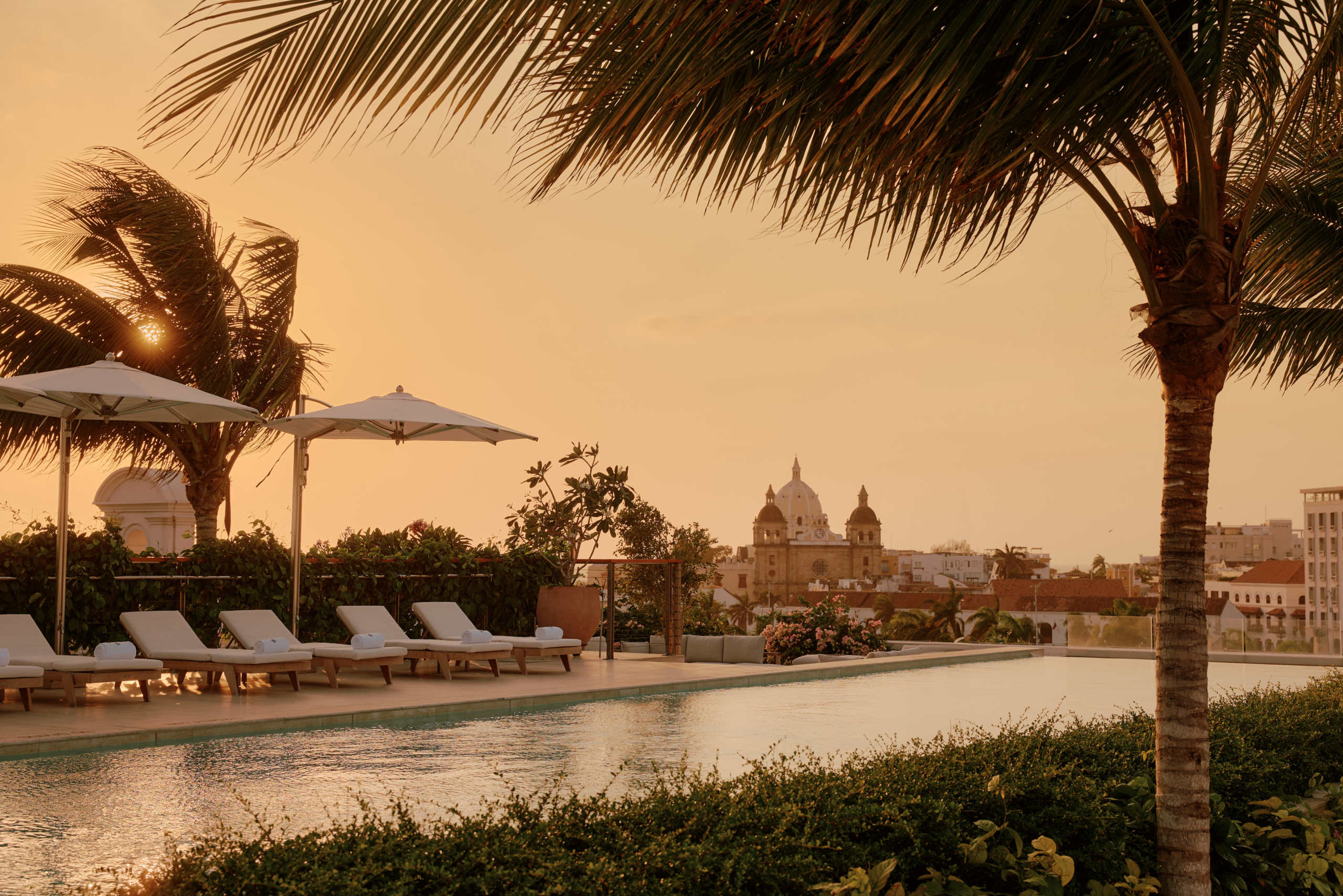 Four Seasons Hotel Cartagena rooftop pool at sunset, with sun loungers and palm trees framing a view over the historic domes and terracotta rooftops of Cartagena's walled city, Colombia.