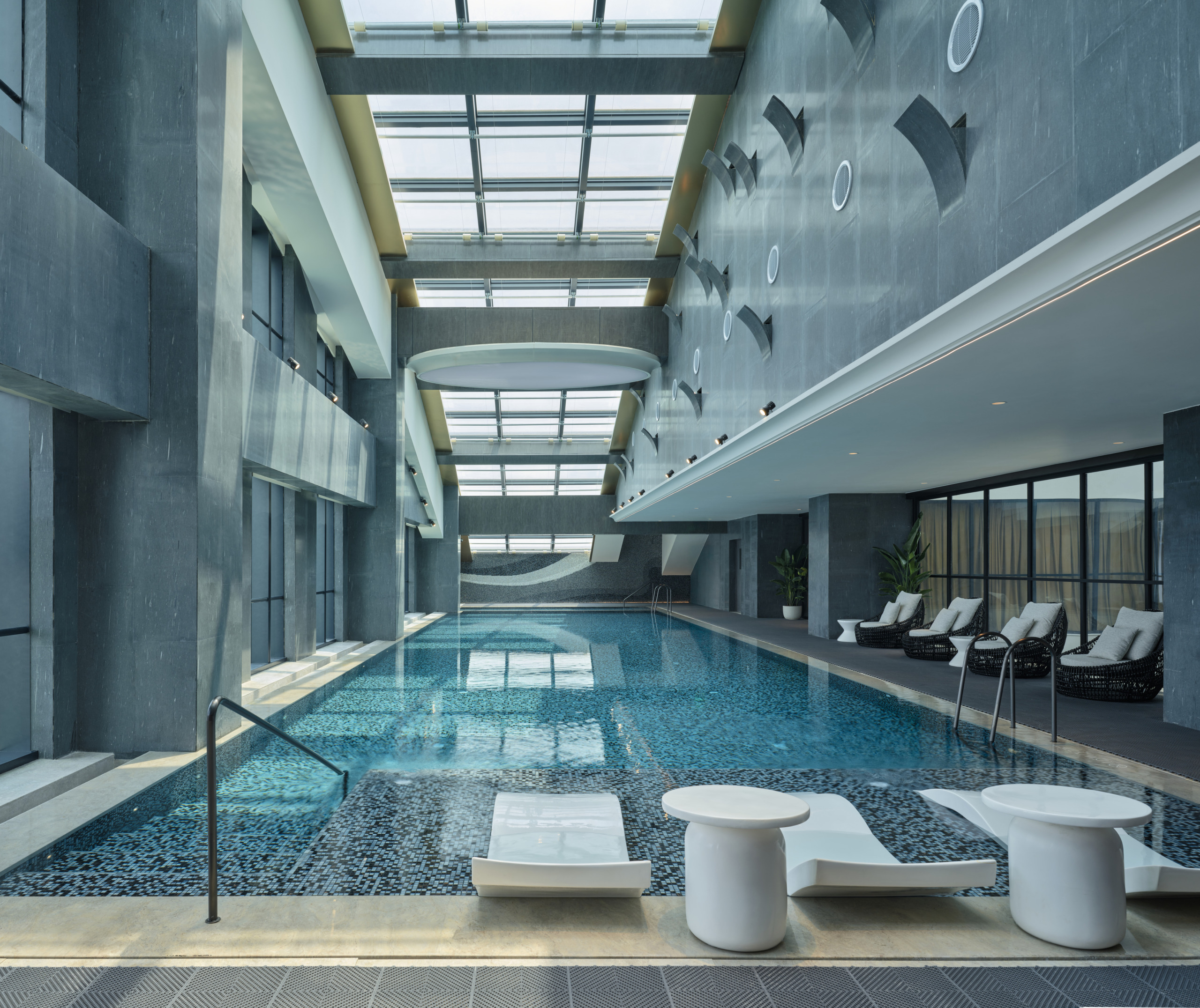 Hotel Indigo Zhuhai indoor lap pool with skylight ceiling, sculptural wall art, white poolside loungers, and rattan sun chairs.