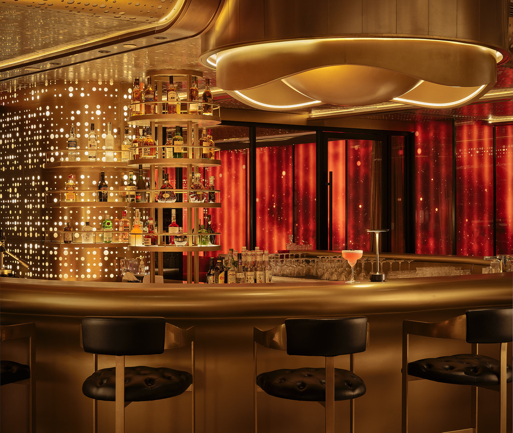 Hotel Indigo Zhuhai rooftop bar counter detail with tiered gold spirit shelves, perforated brass backlit wall, and red draped curtains beyond.