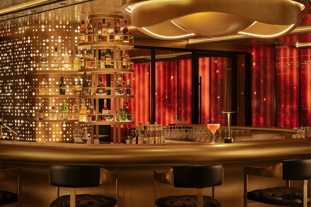 Hotel Indigo Zhuhai rooftop bar counter detail with tiered gold spirit shelves, perforated brass backlit wall, and red draped curtains beyond.