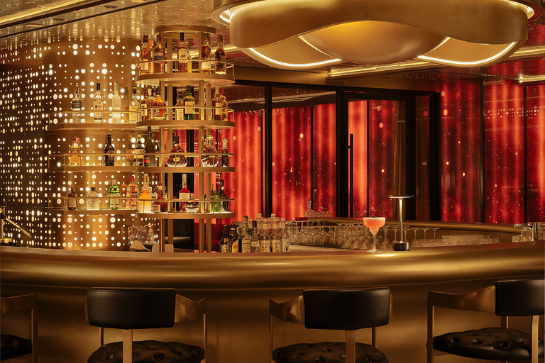 Hotel Indigo Zhuhai rooftop bar counter detail with tiered gold spirit shelves, perforated brass backlit wall, and red draped curtains beyond.