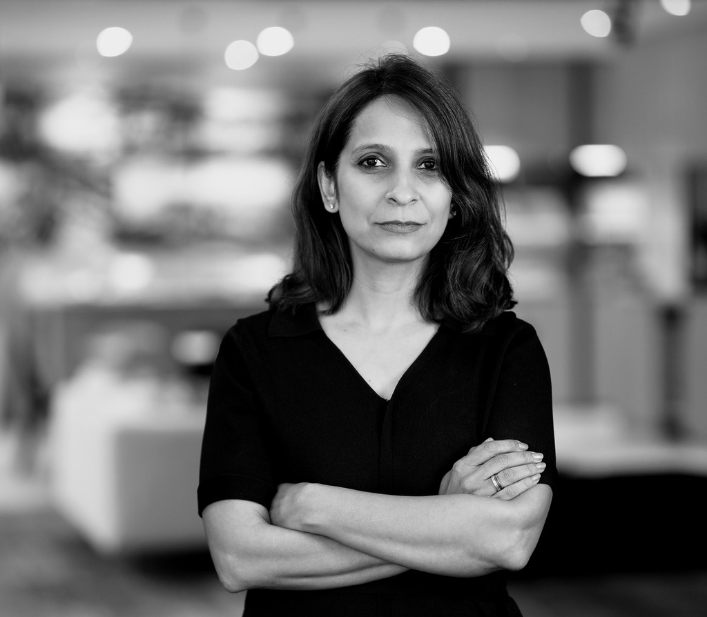 A professional black and white portrait of Bhavya Kukrety, Managing Director of WATG and Wimberly Interiors&rsquo; APAC office. She stands with her arms crossed, wearing a dark V-neck top, against a softly blurred studio background with warm bokeh highlights.