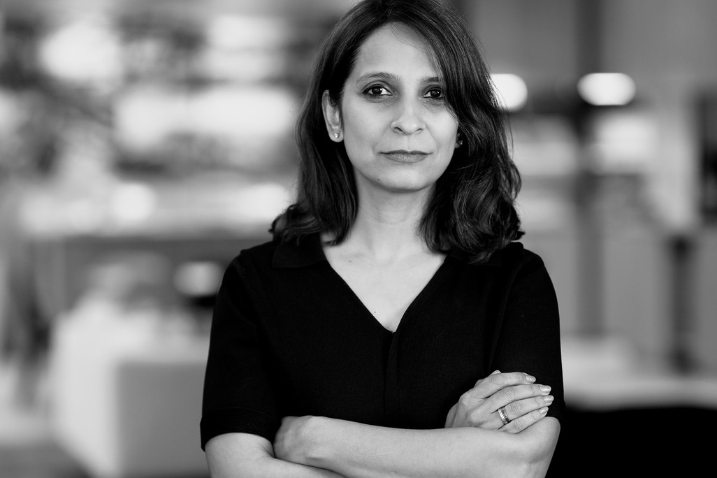 A professional black and white portrait of Bhavya Kukrety, Managing Director of WATG and Wimberly Interiors&rsquo; APAC office. She stands with her arms crossed, wearing a dark V-neck top, against a softly blurred studio background with warm bokeh highlights.