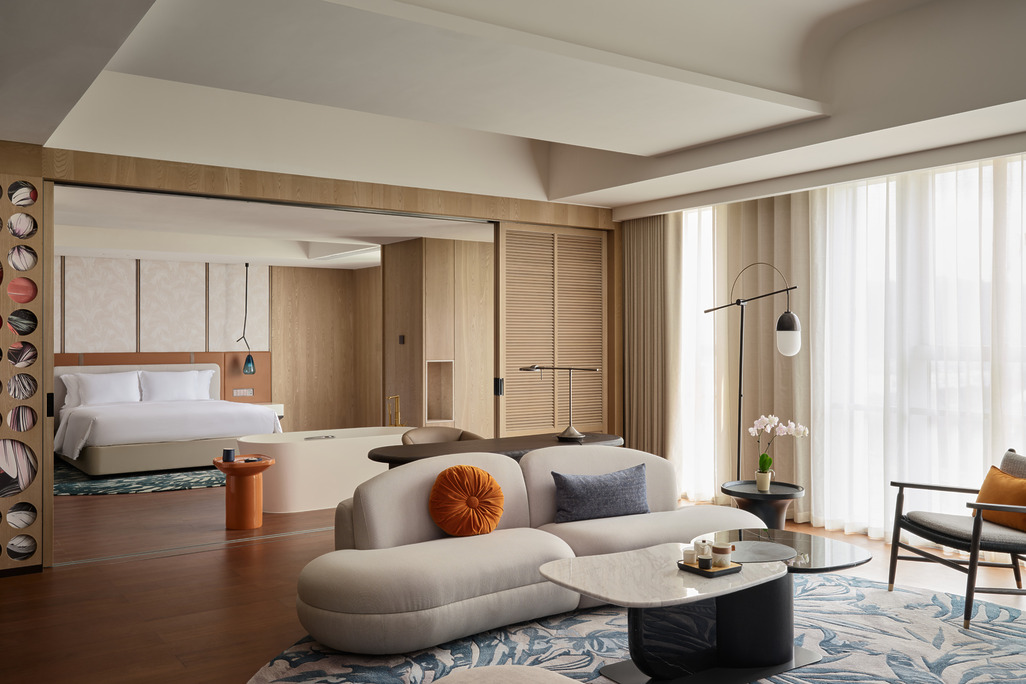 Hotel Indigo Zhuhai suite with curved cream sofa, botanical-print rug, marble coffee table, and sliding partition opening to a king bedroom with warm wood paneling.