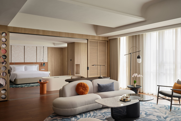 Hotel Indigo Zhuhai suite with curved cream sofa, botanical-print rug, marble coffee table, and sliding partition opening to a king bedroom with warm wood paneling.