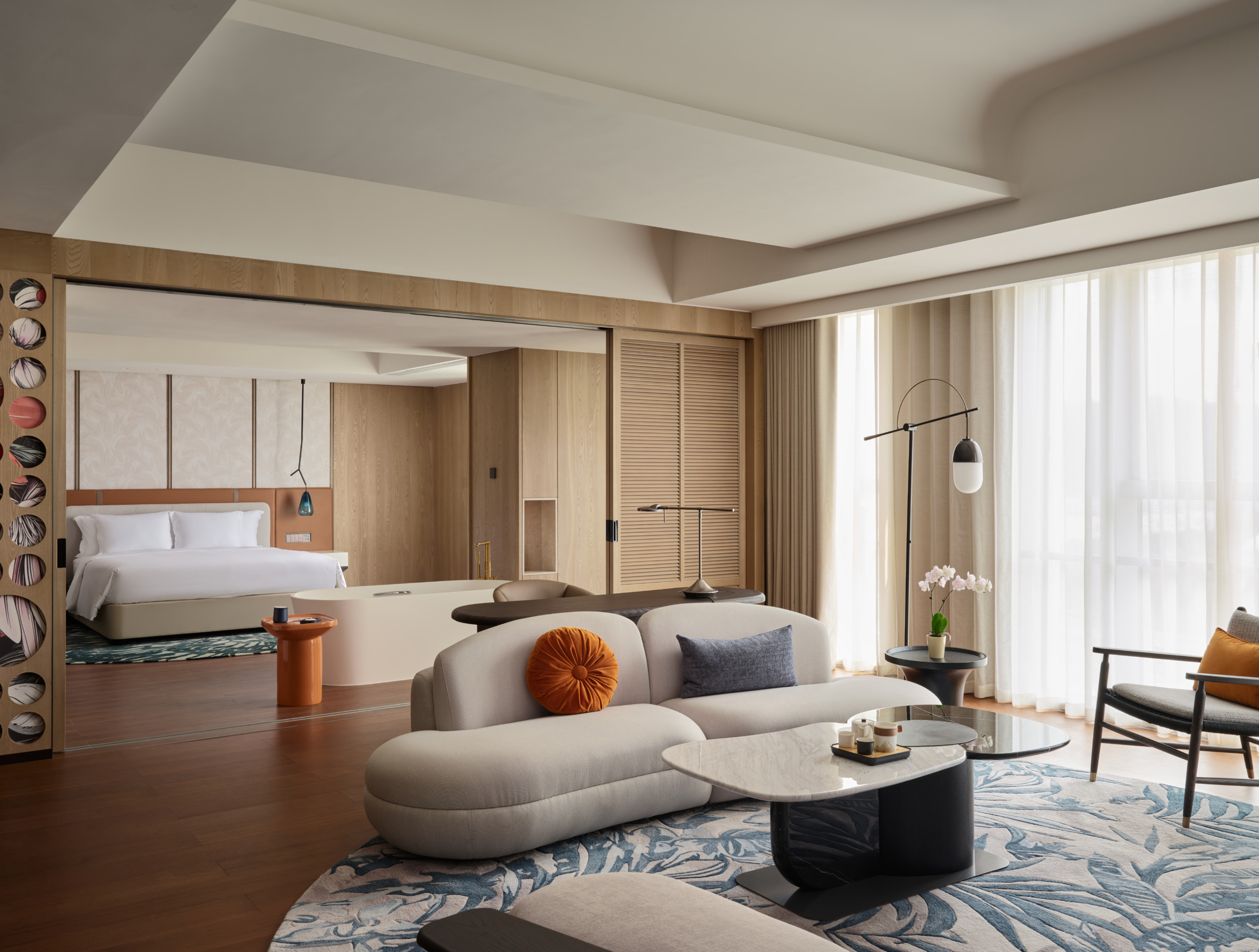 Hotel Indigo Zhuhai suite with curved cream sofa, botanical-print rug, marble coffee table, and sliding partition opening to a king bedroom with warm wood paneling.