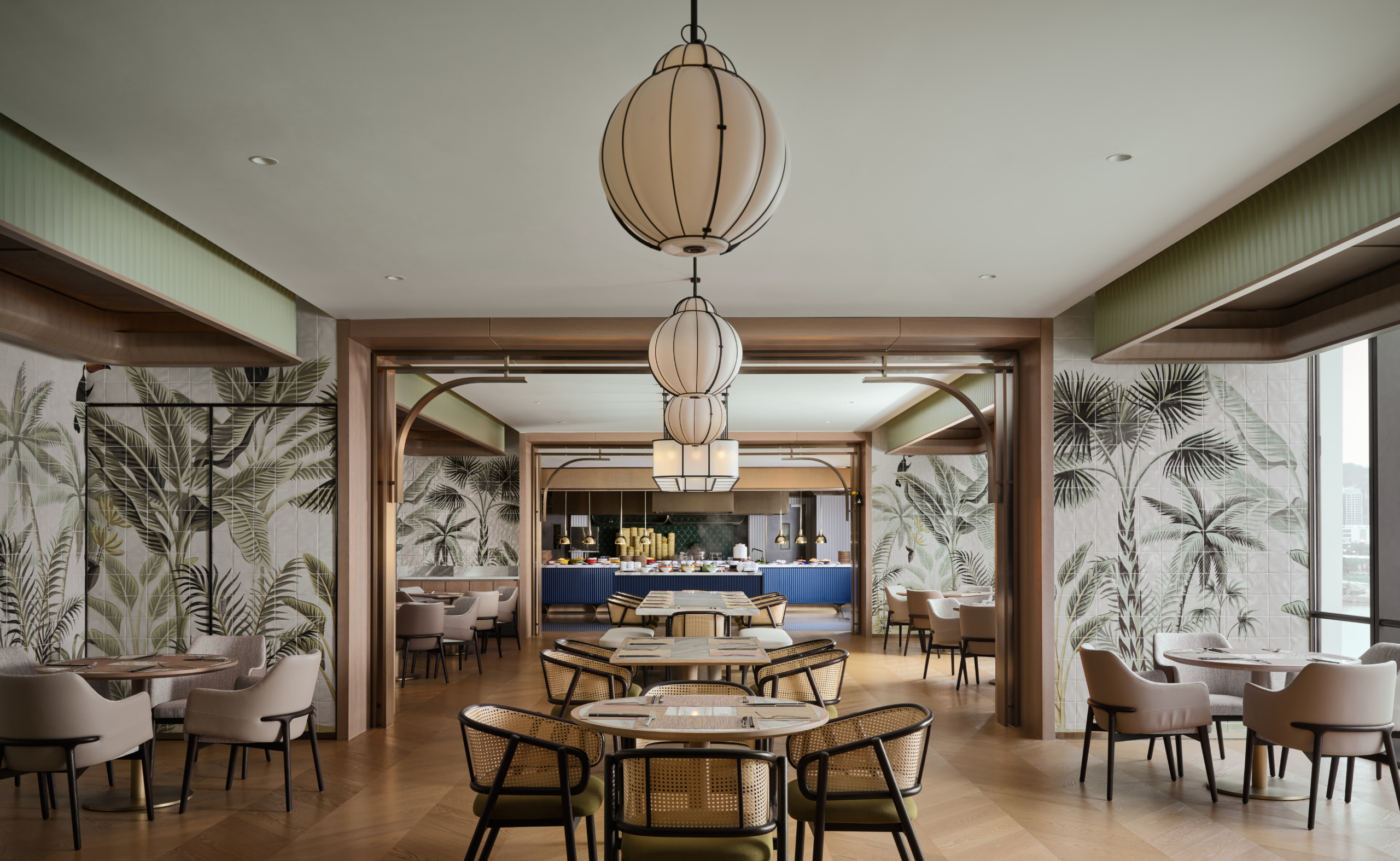 Hotel restaurant with rattan dining chairs, lantern pendants, tropical mural wallpaper, and a blue-fronted buffet counter beyond.