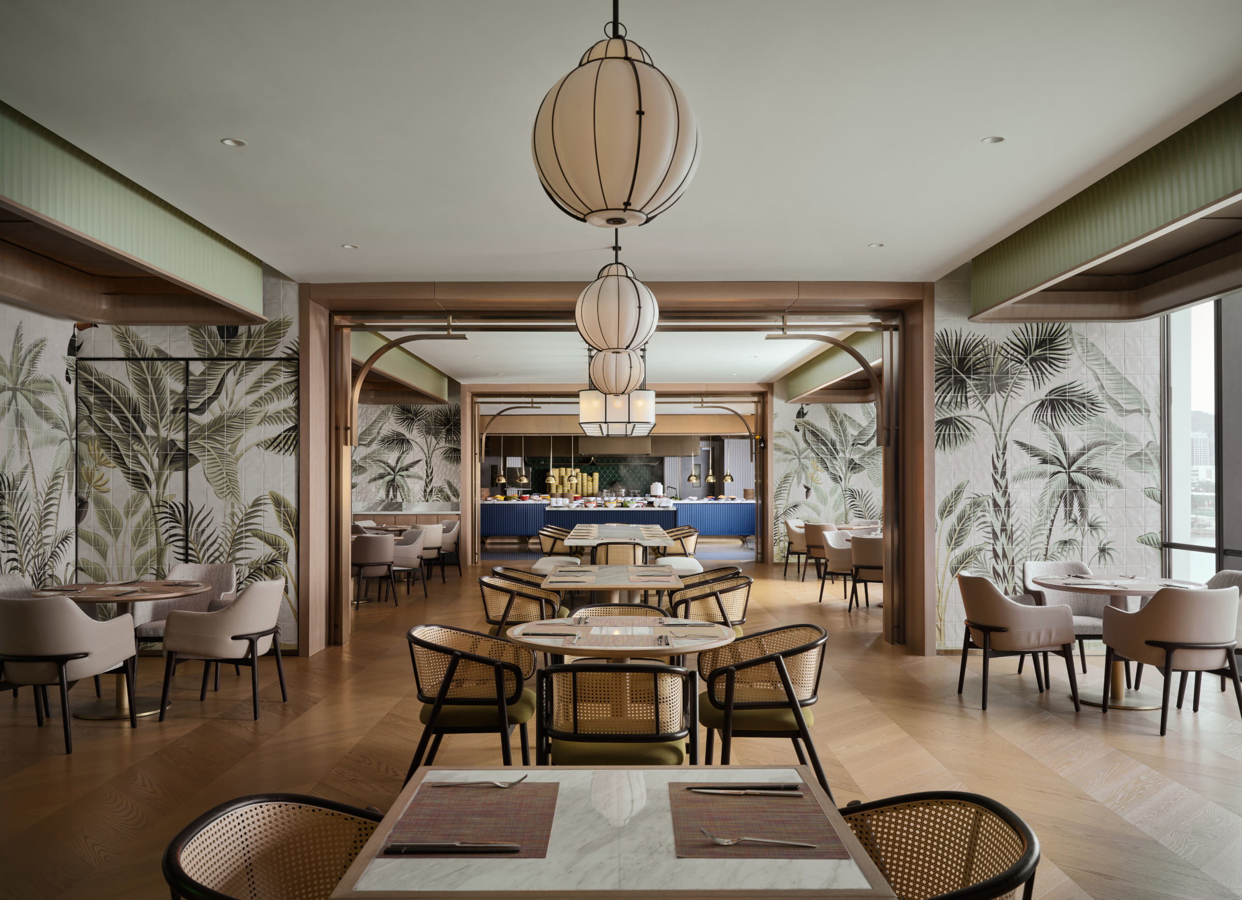 Hotel restaurant with rattan dining chairs, lantern pendants, tropical mural wallpaper, and a blue-fronted buffet counter beyond.