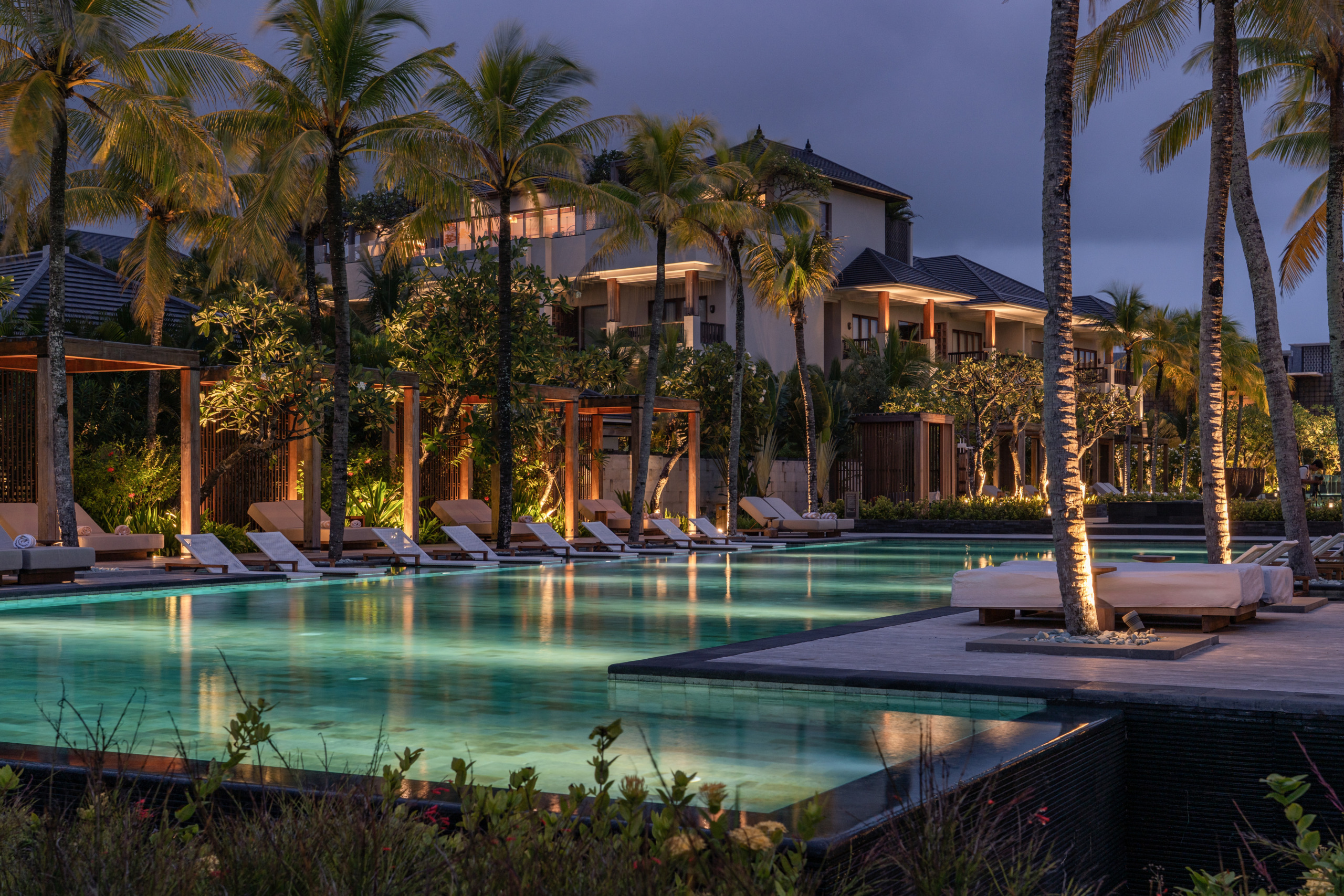 Dusk view of the main pool at The Regent Bali, Canggu, with glowing turquoise water, sun loungers beneath timber pergolas, tall palm trees, and the resort's Balinese-influenced architecture lit against a deep blue evening sky.