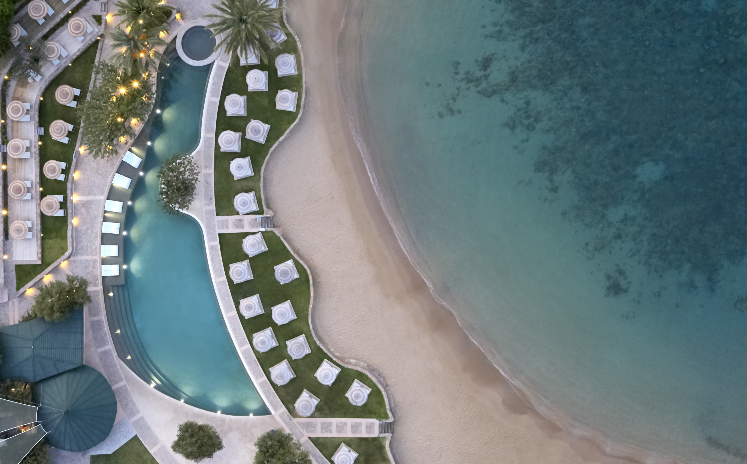 Aerial view of Porto Elounda resort, Crete, showing freeform pool with warm lighting, white sun loungers on manicured lawns, and sandy beach meeting turquoise sea at dusk.