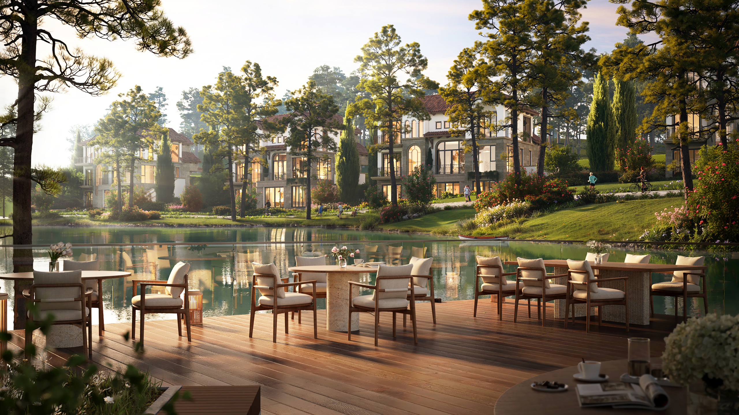 Golden-hour render of a lakeside timber deck at Thanh Xuan Valley, set with outdoor dining tables overlooking the water toward the Residence Villas nestled among pine trees, with residents walking, jogging, and cycling along the shoreline path.