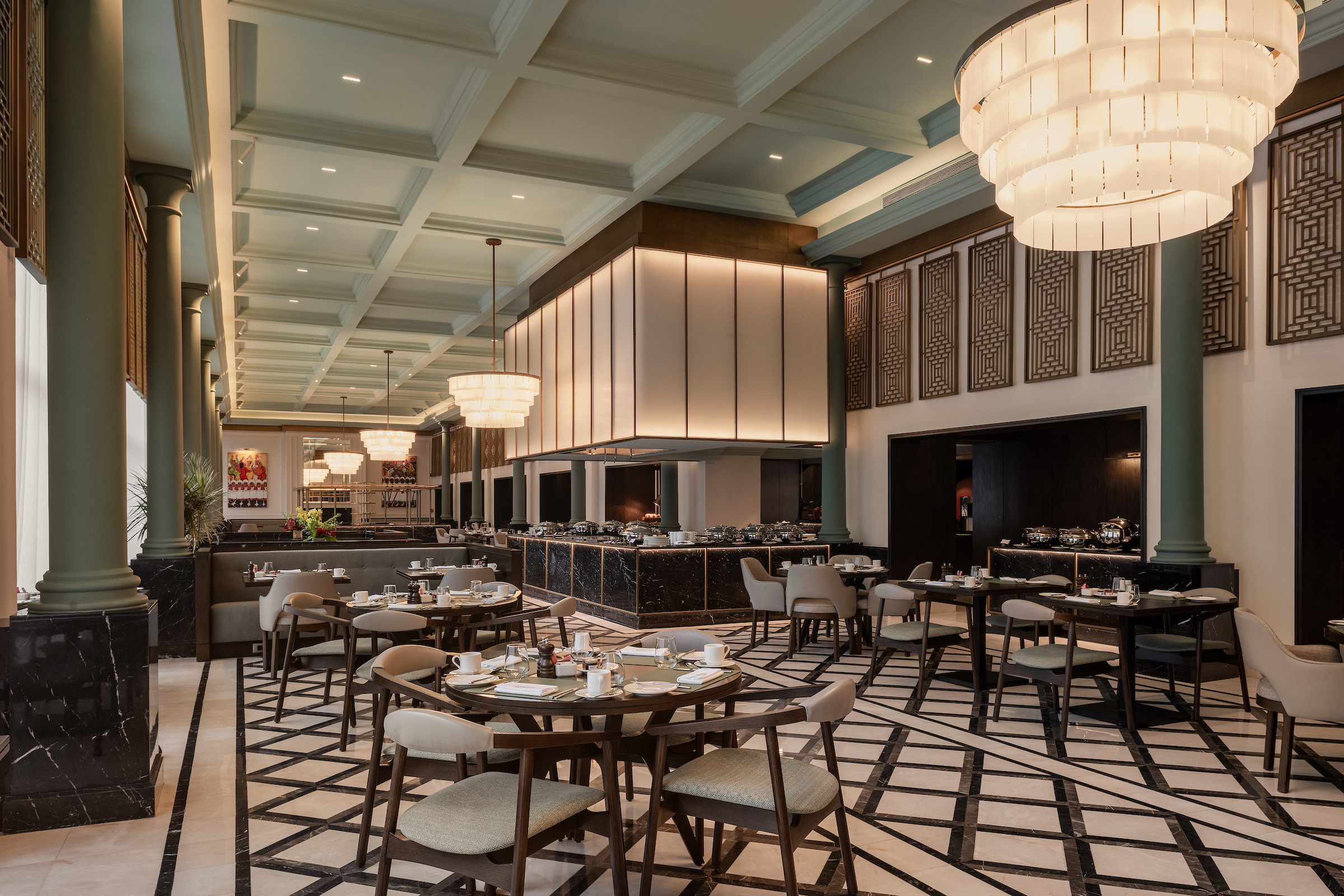Arola restaurant at Giza Palace featuring a coffered ceiling, layered brass-and-glass pendant chandeliers, dark green marble columns, and geometric wood-paneled walls. Round dining tables with taupe upholstered chairs sit atop a black-and-white marble mosaic floor, with a central illuminated buffet counter and chafing dishes visible in the background.