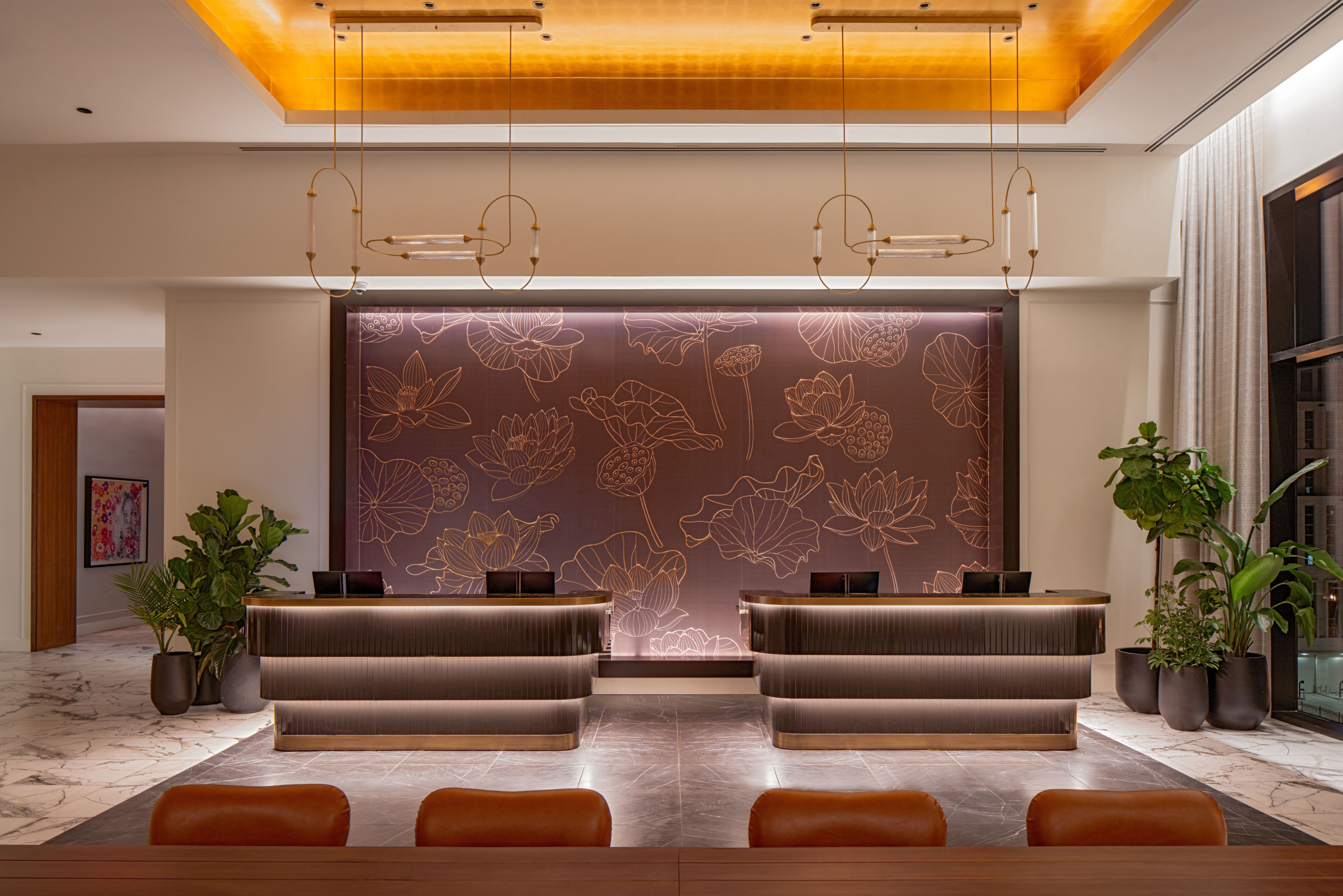 Hotel front desk at Tempo by Hilton Nashville Downtown, featuring twin reception counters in dark wood with brass trim, set against a backlit mauve wall illustrated with a gold lotus flower motif. Sculptural brass pendant lights hang above, and fiddle-leaf fig plants flank the marble-floored lobby. Sunbelt hospitality design.
