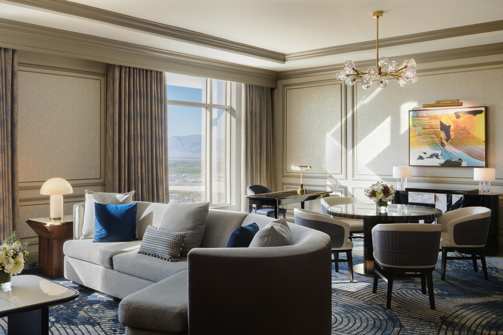 Elegant living room in a Venetian Tower Suite featuring a grey curved sectional sofa with navy and patterned throw pillows, a round dining table with upholstered chairs, brass chandelier, and floor-to-ceiling windows overlooking the Las Vegas skyline and mountains.